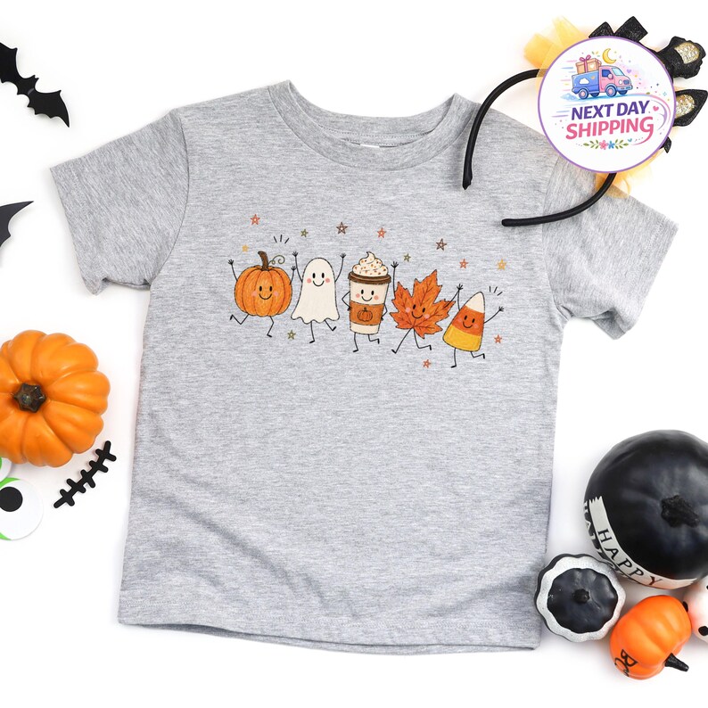 May include: A heather gray short-sleeve t-shirt featuring a graphic print of a smiling pumpkin, a ghost, a coffee cup, an autumn leaf, and a piece of candy corn, all arranged in a row with small star accents scattered around the characters.
