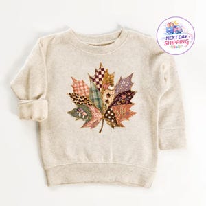 Puede incluir: Una sudadera beige de manga larga con un gran gr&aacute;fico central de una hoja de arce compuesta por varios parches estampados, incluyendo cuadros, estampado de leopardo, lunares y flores, con peque&ntilde;as ilustraciones de setas y bellotas.
