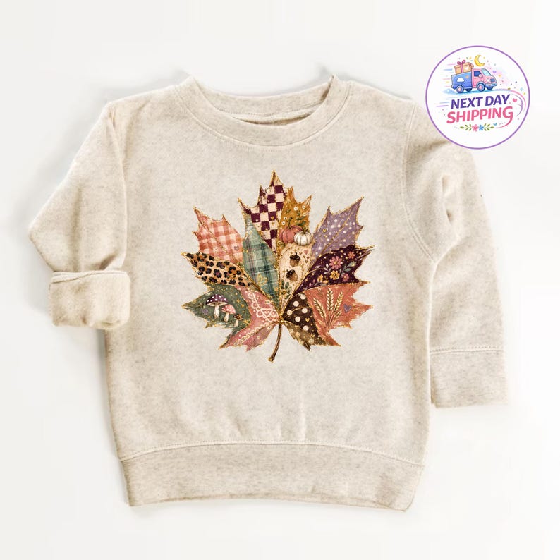 Puede incluir: Una sudadera beige de manga larga con un gran gr&aacute;fico central de una hoja de arce compuesta por varios parches estampados, incluyendo cuadros, estampado de leopardo, lunares y flores, con peque&ntilde;as ilustraciones de setas y bellotas.