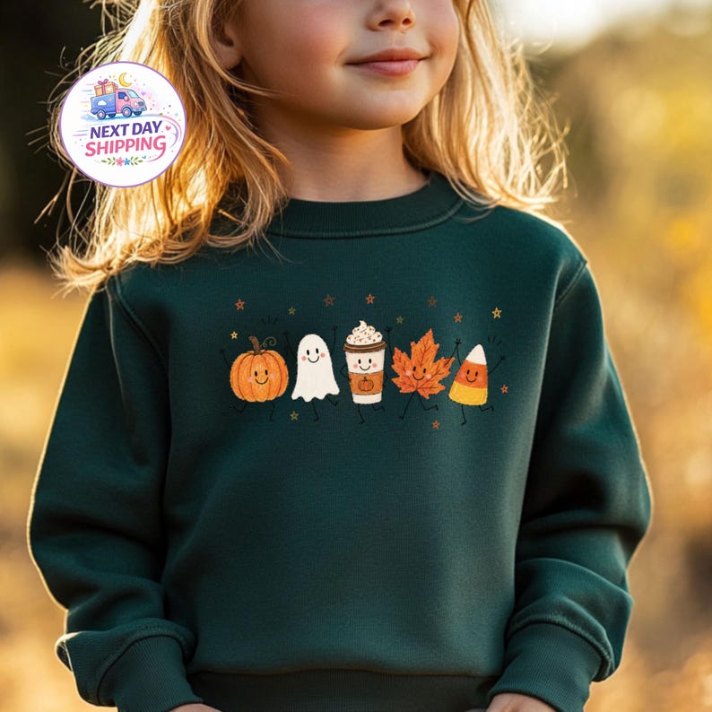 May include: A dark green long-sleeve sweatshirt featuring a graphic print of five smiling autumn-themed characters: a pumpkin, a ghost, a coffee cup, an orange maple leaf, and a piece of candy corn, all accented with small scattered stars.