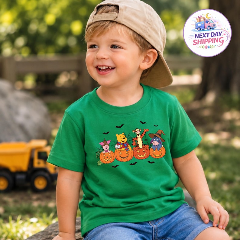 May include: A bright green short-sleeve t-shirt featuring a graphic print of Piglet, Winnie the Pooh, Tigger, and Eeyore sitting inside carved orange jack-o'-lantern pumpkins, accented with black silhouettes of flying bats and spider webs.