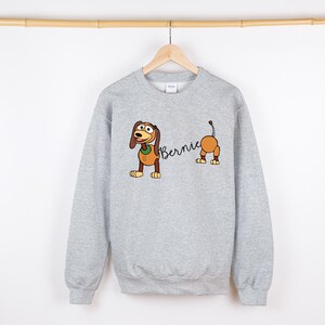 Op de afbeelding: Een lichtgrijs sweatshirt met een cartoon teckel ontwerp. De teckel is bruin en bruin met de naam "Bernie" in een scriptlettertype. De hond is in twee delen, voor- en achterkant gescheiden.