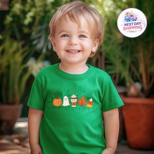 May include: A bright green short-sleeved t-shirt featuring a graphic print of a smiling pumpkin, a ghost, a coffee cup, an autumn leaf, and a piece of candy corn, each with small stick arms and legs, arranged in a horizontal row across the chest.