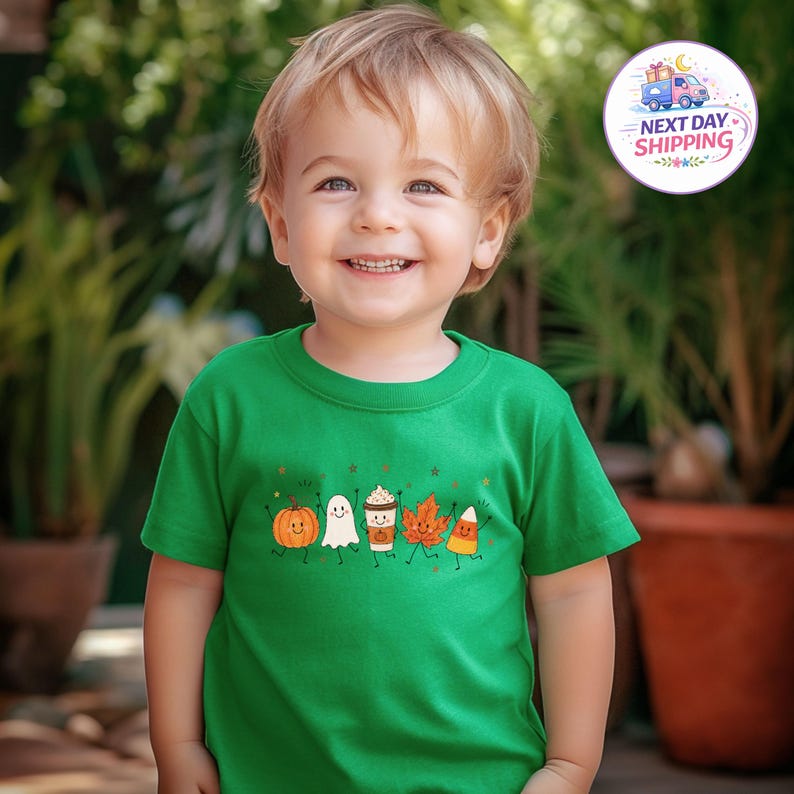 May include: A bright green short-sleeved t-shirt featuring a graphic print of a smiling pumpkin, a ghost, a coffee cup, an autumn leaf, and a piece of candy corn, each with small stick arms and legs, arranged in a horizontal row across the chest.