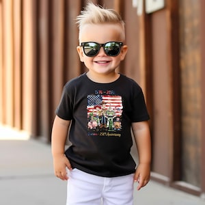 K&ouml;nnte beinhalten: Ein schwarzes T-Shirt mit einer Grafik der amerikanischen Flagge und Toy Story-Charakteren. Das Shirt zeigt den Text "1776-2026" und "America - 250th Anniversary". Das Kind tr&auml;gt eine Sonnenbrille und wei&szlig;e Shorts.