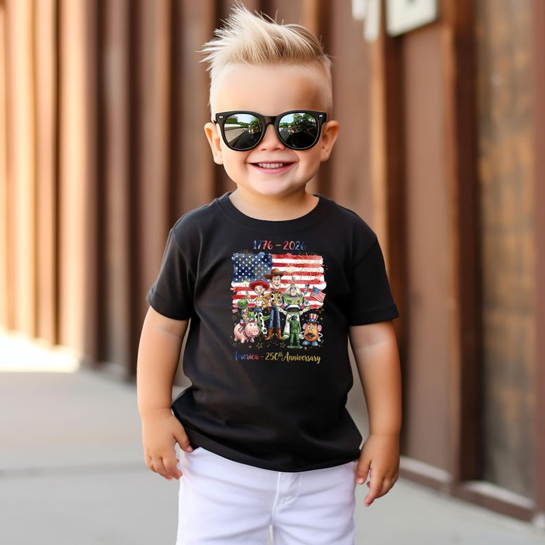 K&ouml;nnte beinhalten: Ein schwarzes T-Shirt mit einer Grafik der amerikanischen Flagge und Toy Story-Charakteren. Das Shirt zeigt den Text "1776-2026" und "America - 250th Anniversary". Das Kind tr&auml;gt eine Sonnenbrille und wei&szlig;e Shorts.