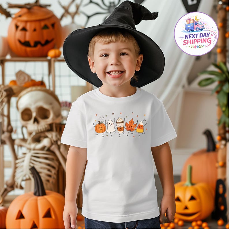 May include: A white short-sleeved t-shirt featuring a graphic print of five smiling autumn-themed characters: a pumpkin, a ghost, a coffee cup, an orange maple leaf, and a piece of candy corn, all arranged in a row on the chest of the garment.