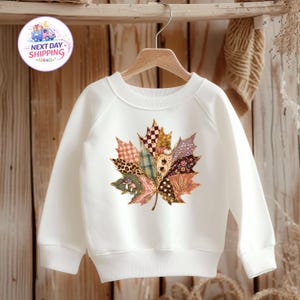Puede incluir: Una sudadera blanca de manga larga con un gran gr&aacute;fico central de una hoja de arce compuesta por varios segmentos estampados, incluyendo cuadros, estampado de leopardo, lunares y dise&ntilde;os florales en tonos marr&oacute;n, naranja y crema.