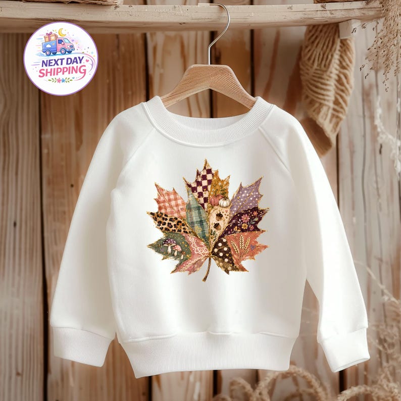 Puede incluir: Una sudadera blanca de manga larga con un gran gr&aacute;fico central de una hoja de arce compuesta por varios segmentos estampados, incluyendo cuadros, estampado de leopardo, lunares y dise&ntilde;os florales en tonos marr&oacute;n, naranja y crema.