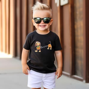 Op de afbeelding: Zwart T-shirt met een cartoon hond ontwerp en de naam "Bernie". De hond is bruin en bruin met een groene kraag. Het shirt wordt gedragen met een witte korte broek. Het kind draagt een zwarte zonnebril.