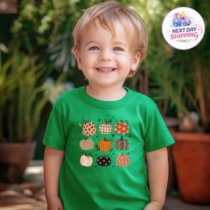 May include: A bright green short-sleeved t-shirt featuring a graphic print of nine pumpkins in various patterns, including leopard print, plaid, floral, and solid orange. The pumpkins are arranged in a three-by-three grid on the chest area.