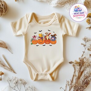 May include: A cream-coloured short-sleeve baby bodysuit featuring a graphic print of Piglet, Winnie the Pooh, Tigger, and Eeyore sitting inside orange jack-o'-lantern pumpkins, surrounded by black bats and spiderwebs on the chest area.