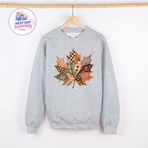 Puede incluir: Una sudadera gris claro de cuello redondo con un gran gr&aacute;fico central de una hoja de arce compuesta por varios patrones de patchwork, incluyendo cuadros, estampado de leopardo, lunares y dise&ntilde;os florales en tonos marr&oacute;n, naranja y crema.