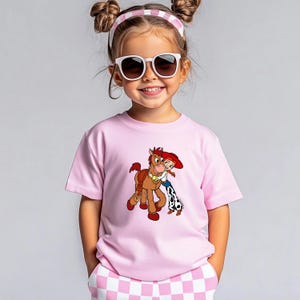 Puede incluir: Una camiseta rosa claro con un dibujo animado de un caballo y una vaquera. La camiseta de manga corta la lleva un ni&ntilde;o peque&ntilde;o. El ni&ntilde;o lleva gafas de sol y una diadema rosa y blanca.