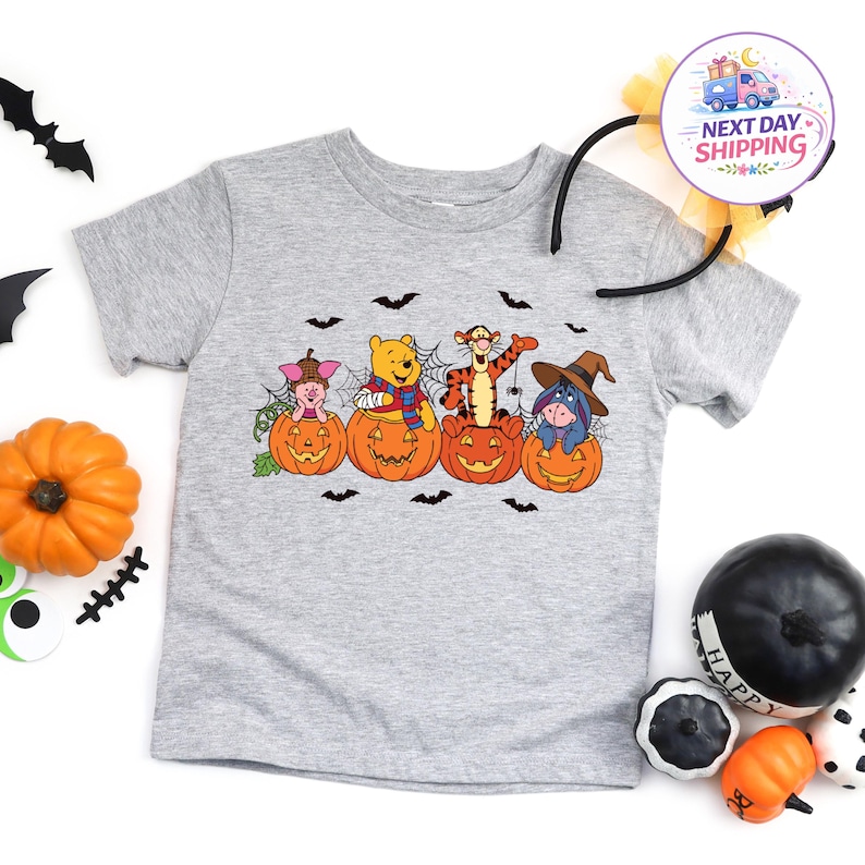 May include: A heather grey short-sleeved t-shirt featuring a graphic print of Piglet, Winnie the Pooh, Tigger, and Eeyore sitting inside orange jack-o'-lantern pumpkins, surrounded by black bat silhouettes and spider web accents.