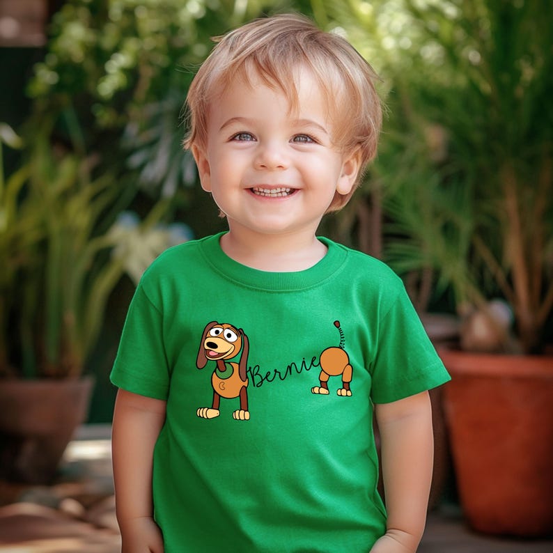 Op de afbeelding: Een groen t-shirt met een cartoon hond ontwerp en de naam "Bernie". De hond is bruin en beige. Het t-shirt wordt gedragen door een jong kind. De achtergrond is wazig met groen gebladerte.