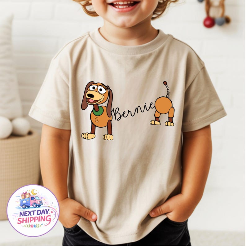 Op de afbeelding: Beige T-shirt met korte mouwen met een cartoonhondontwerp en de naam "Bernie". De hond is bruin, beige en wit met een groene kraag. De staart van de hond is ook zichtbaar.
