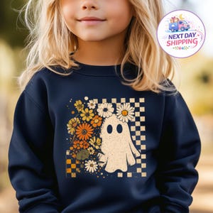 May include: A navy blue long-sleeve sweatshirt featuring a graphic print of a white ghost surrounded by orange, yellow, and white flowers. The design is set against a checkered background pattern, creating a contrast with the dark fabric of the shirt.