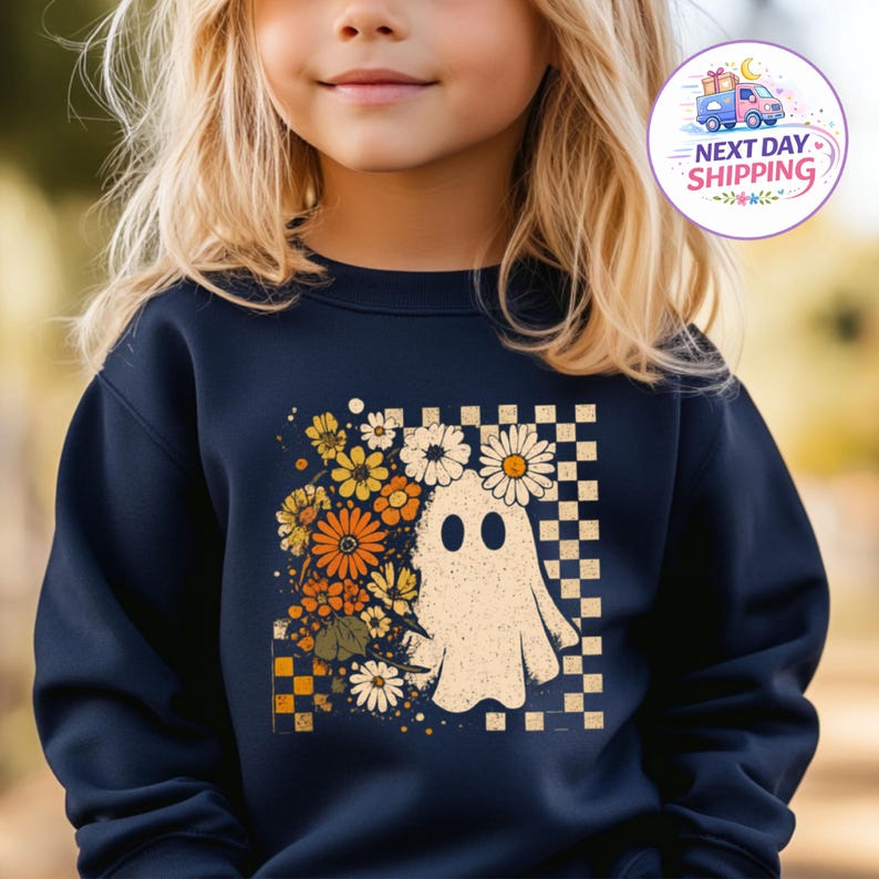 May include: A navy blue long-sleeve sweatshirt featuring a graphic print of a white ghost surrounded by orange, yellow, and white flowers. The design is set against a checkered background pattern, creating a contrast with the dark fabric of the shirt.