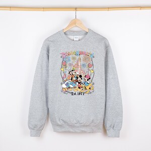 Op de afbeelding: Een lichtgrijze sweatshirt met een kleurrijke afbeelding van Disney-personages, waaronder Mickey Mouse, Minnie Mouse, Donald Duck en Goofy, voor een kasteel. De tekst "Disneyworld" en "Est. 1971" zijn ook afgebeeld.