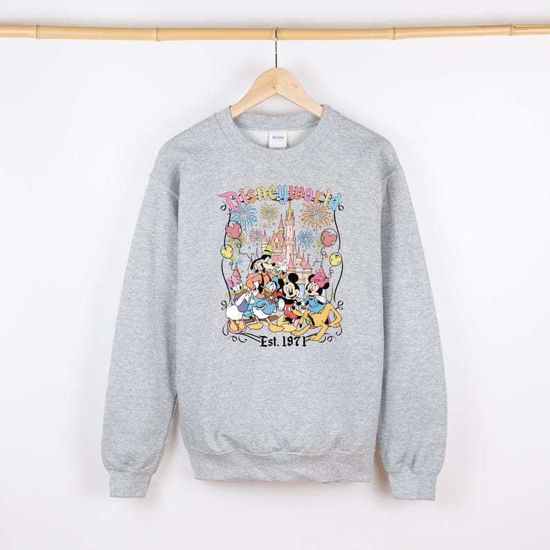 Op de afbeelding: Een lichtgrijze sweatshirt met een kleurrijke afbeelding van Disney-personages, waaronder Mickey Mouse, Minnie Mouse, Donald Duck en Goofy, voor een kasteel. De tekst "Disneyworld" en "Est. 1971" zijn ook afgebeeld.
