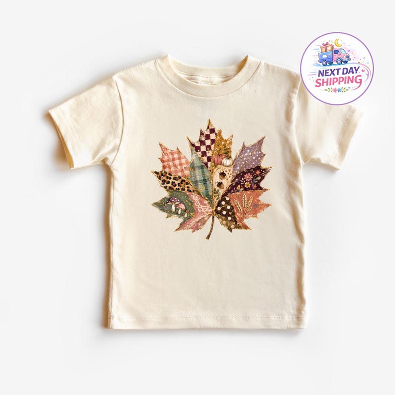 Puede incluir: Una camiseta de manga corta color crema con un gran gr&aacute;fico central de una hoja de arce compuesta por varios segmentos estampados, incluyendo estampado de leopardo, cuadros, lunares, setas y motivos florales en tonos oto&ntilde;ales.