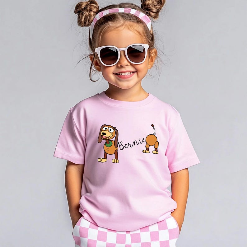 Op de afbeelding: Lichtroze T-shirt met een cartoon hond ontwerp en de naam "Bernie". De hond is bruin en bruin met een groene kraag. Het kind draagt een zonnebril en een roze en witte hoofdband.