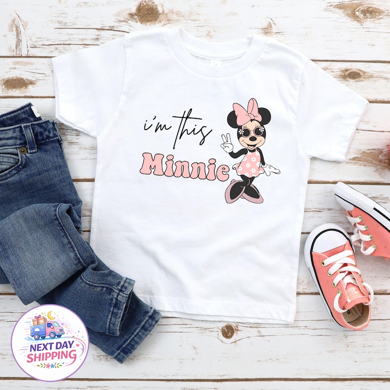 May include: White t-shirt featuring a Minnie Mouse graphic with the text "I'm this Minnie". The design includes Minnie Mouse wearing sunglasses and a pink bow. The shirt is paired with blue jeans and pink sneakers.