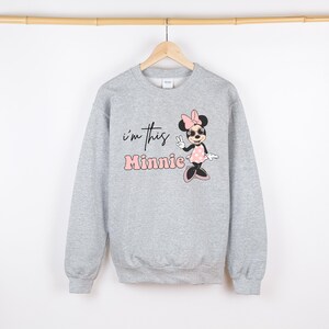 May include: A light gray sweatshirt featuring a Minnie Mouse graphic. The graphic includes the text "I'm this Minnie" in black and pink. Minnie Mouse is depicted wearing sunglasses, a pink bow, and a polka dot dress.