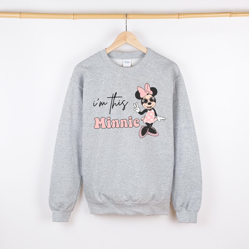 May include: A light gray sweatshirt featuring a Minnie Mouse graphic. The graphic includes the text "I'm this Minnie" in black and pink. Minnie Mouse is depicted wearing sunglasses, a pink bow, and a polka dot dress.