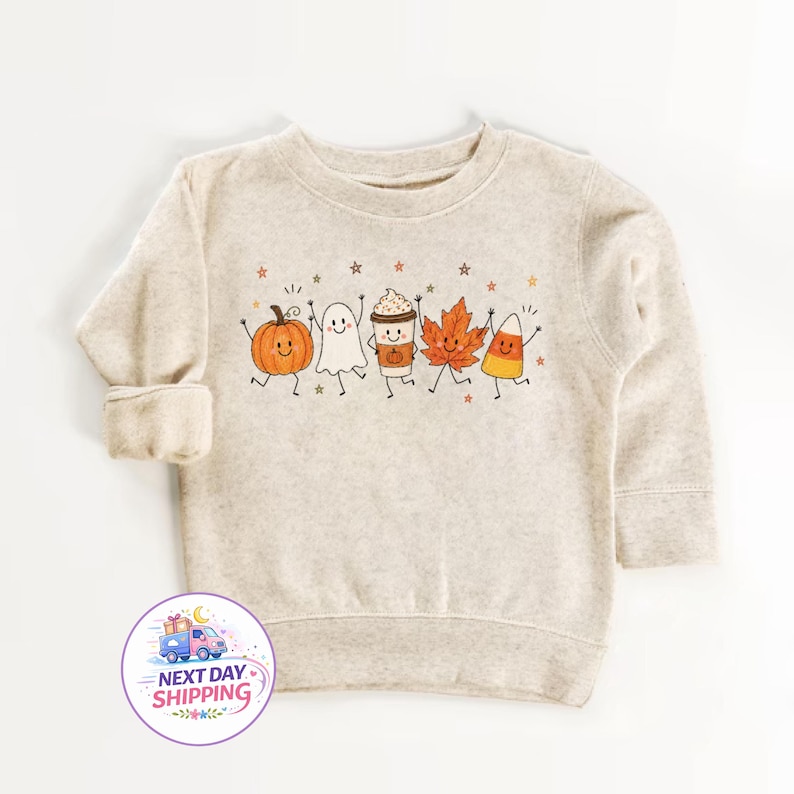 May include: A heathered beige long-sleeve sweatshirt featuring a central graphic of five cartoon characters: an orange pumpkin, a white ghost, a coffee cup, an orange maple leaf, and a candy corn, all with smiling faces and small stars.