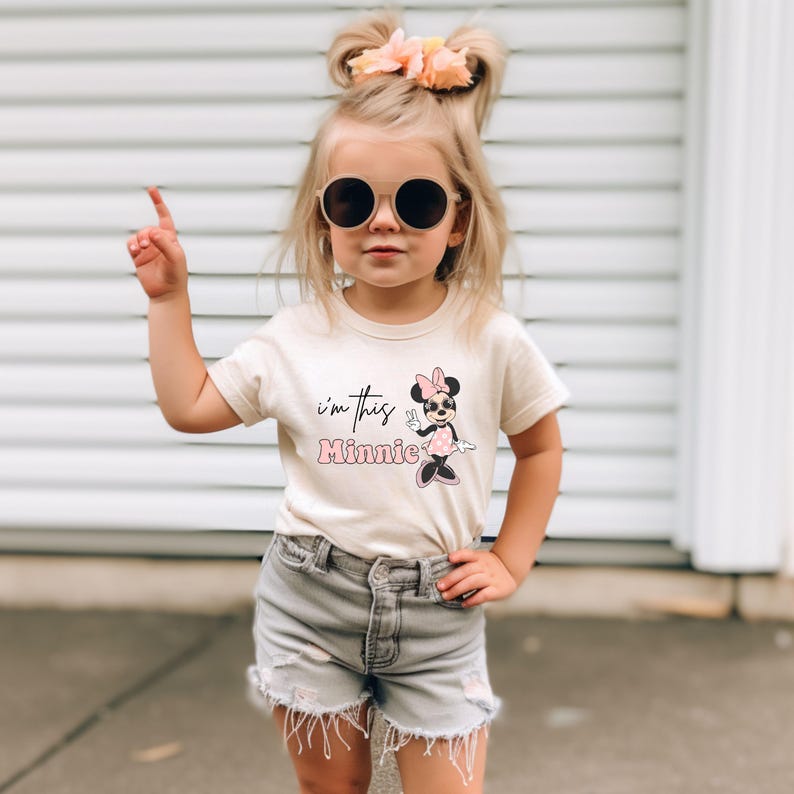 May include: A young child wearing a beige t-shirt with the text "I'm this Minnie" and a Minnie Mouse graphic. The child is also wearing gray shorts and round sunglasses. The child is pointing upwards with one finger.