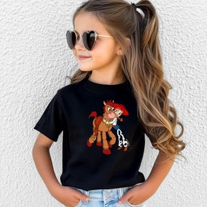 Puede incluir: Camiseta negra con un dibujo animado de un caballo marr&oacute;n y una ni&ntilde;a con el pelo rojo. La ni&ntilde;a lleva una camisa blanca, un chaleco azul y pantalones con estampado de vaca. La ni&ntilde;a lleva gafas de sol con forma de coraz&oacute;n.