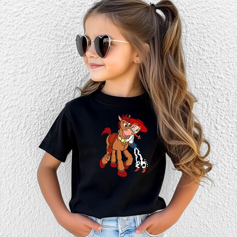 Puede incluir: Camiseta negra con un dibujo animado de un caballo marr&oacute;n y una ni&ntilde;a con el pelo rojo. La ni&ntilde;a lleva una camisa blanca, un chaleco azul y pantalones con estampado de vaca. La ni&ntilde;a lleva gafas de sol con forma de coraz&oacute;n.