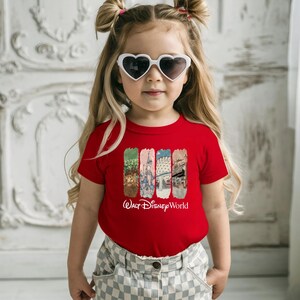Puede incluir: Una ni&ntilde;a lleva una camiseta roja con un gr&aacute;fico colorido de los lugares emblem&aacute;ticos de Walt Disney World. La camiseta presenta una serie de rect&aacute;ngulos verticales con im&aacute;genes de un castillo, un puente y otras atracciones. Las palabras "Walt Disney World" est&aacute;n impresas en blanco.