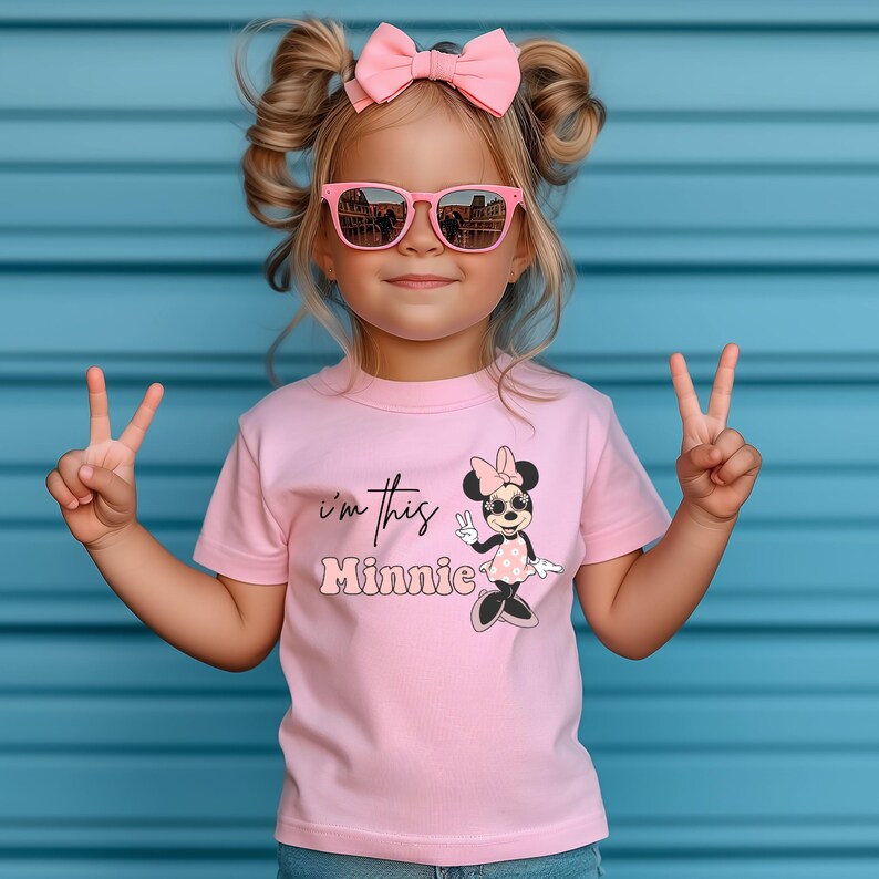 May include: A young child wearing a pink t-shirt with the text "I'm this Minnie" and a Minnie Mouse graphic. The child is also wearing pink sunglasses and a pink bow. The child is giving the peace sign.