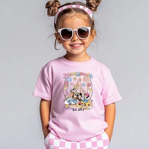 Op de afbeelding: Een kind draagt een roze T-shirt met een Disney World-afbeelding. De afbeelding toont Disney-personages, een kasteel, vuurwerk en de tekst "Disney World Est. 1971". Het kind draagt ook een zonnebril en een roze en witte haarband.