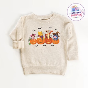 May include: A beige long-sleeve sweatshirt featuring a graphic print of Piglet, Winnie the Pooh, Tigger, and Eeyore, each sitting inside a carved orange jack-o'-lantern pumpkin. The design includes spiderwebs, small black bats, and a spider.