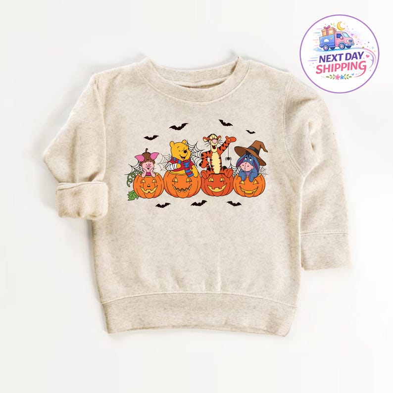 May include: A beige long-sleeve sweatshirt featuring a graphic print of Piglet, Winnie the Pooh, Tigger, and Eeyore, each sitting inside a carved orange jack-o'-lantern pumpkin. The design includes spiderwebs, small black bats, and a spider.