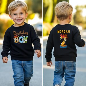 May include: Black sweatshirt with the text "Birthday Boy" in a colorful design. The design includes a cowboy hat and the word "BOY" in a playful font. The back of the sweatshirt has the name "MORGAN" and a number 2 with a cowboy theme.