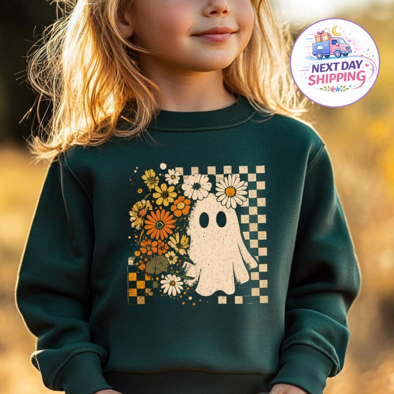 May include: A dark green long-sleeve sweatshirt featuring a graphic print of a white ghost surrounded by orange, yellow, and white flowers. The design includes a checkered pattern background in shades of tan and dark green behind the ghost.