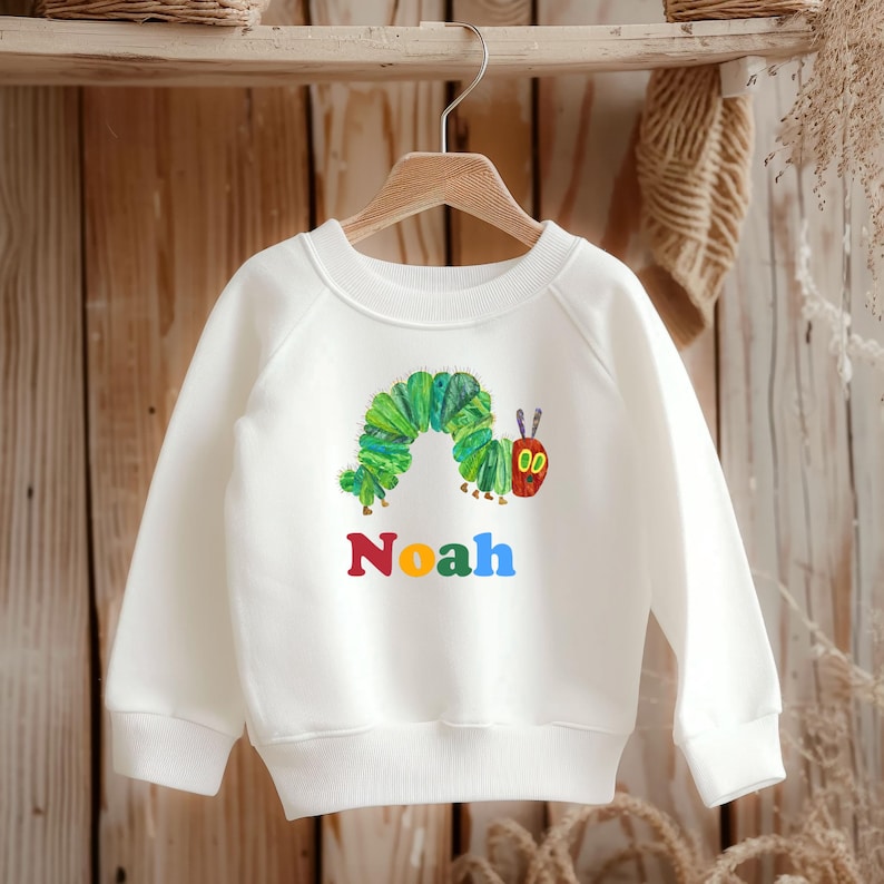 Może przedstawiać: Biała bluza z kolorową ilustracją zielonej gąsienicy z czerwoną twarzą. Imię "Noah" jest wydrukowane w kolorze czerwonym, ż&oacute;łtym i niebieskim poniżej. Bluza wisi na drewnianym wieszaku.