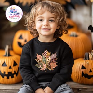 Puede incluir: Una sudadera negra de manga larga con un gran gr&aacute;fico central de hoja de arce compuesto por varios patrones de patchwork, incluyendo cuadros, estampado de leopardo y dise&ntilde;os florales, usada por un ni&ntilde;o de cabello ondulado entre calabazas.