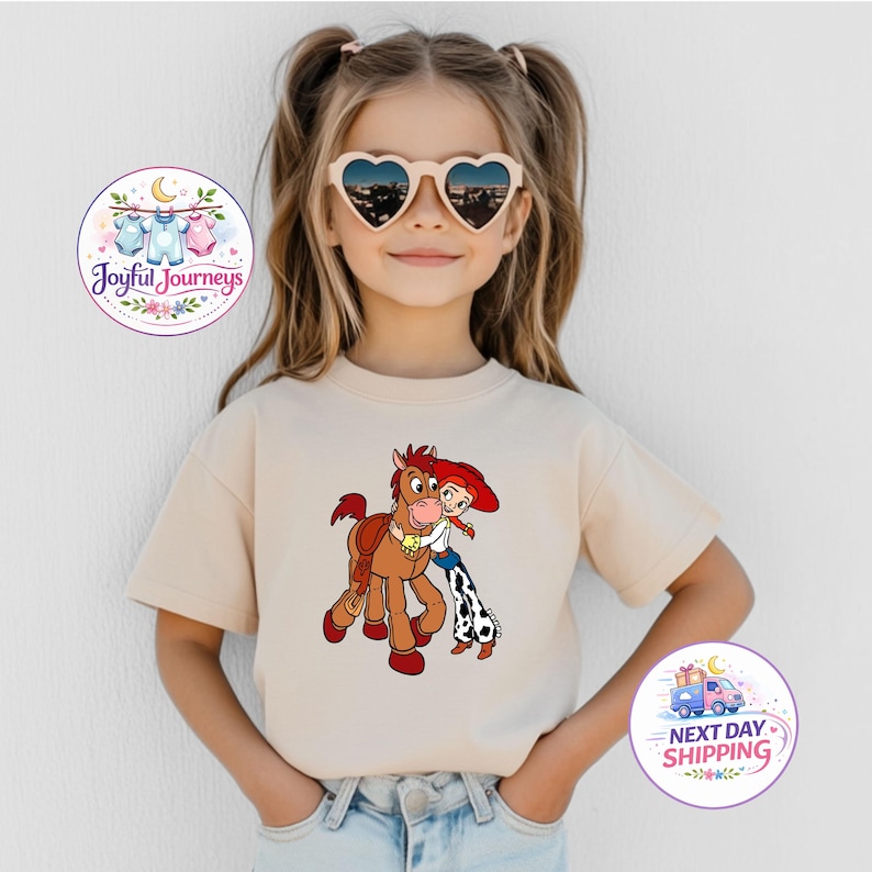 Puede incluir: Una camiseta beige con una ilustraci&oacute;n de dibujos animados de un caballo y una ni&ntilde;a con el pelo rojo. La ni&ntilde;a lleva unos chaps con estampado de vaca. La camiseta la lleva una ni&ntilde;a peque&ntilde;a con gafas de sol en forma de coraz&oacute;n.