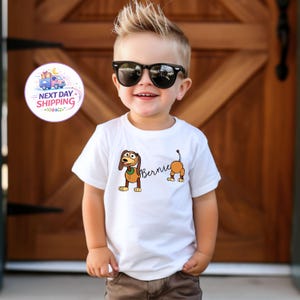 Op de afbeelding: Een wit t-shirt met een cartoon hond ontwerp en de naam "Bernie". De hond is bruin en bruin met een groene kraag. Het t-shirt wordt gedragen door een jong kind.