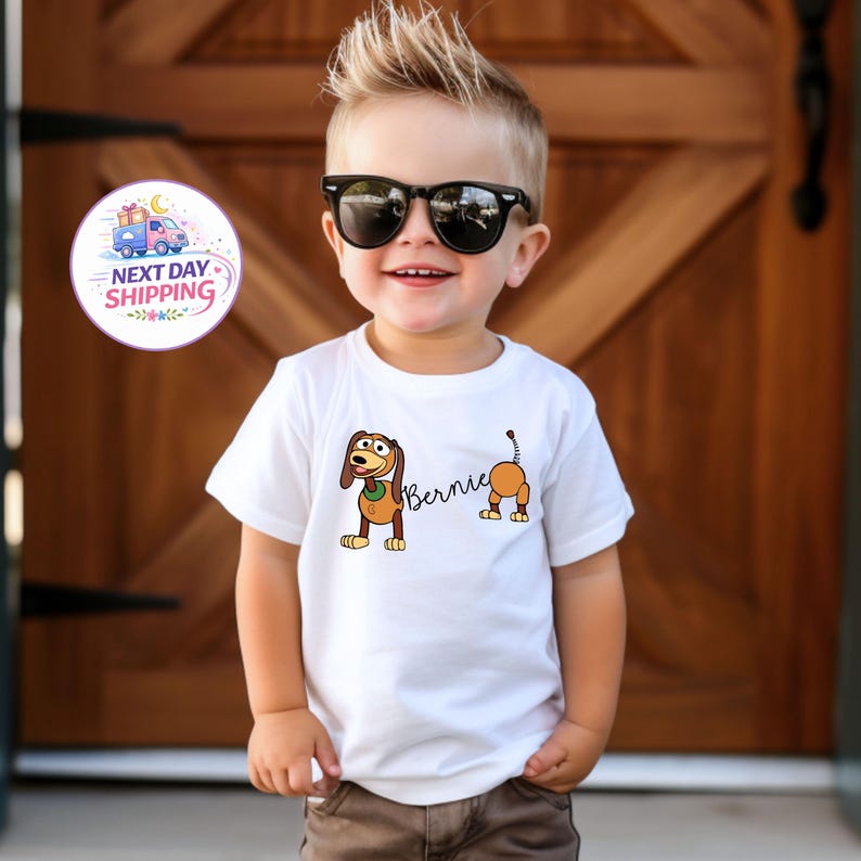 Op de afbeelding: Een wit t-shirt met een cartoon hond ontwerp en de naam "Bernie". De hond is bruin en bruin met een groene kraag. Het t-shirt wordt gedragen door een jong kind.