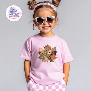 Puede incluir: Una camiseta de manga corta de color rosa claro con un gr&aacute;fico central grande de una hoja de arce compuesta por varios segmentos de tela estampada, incluyendo cuadros, estampado de leopardo y dise&ntilde;os florales, junto a pantalones cortos a cuadros.