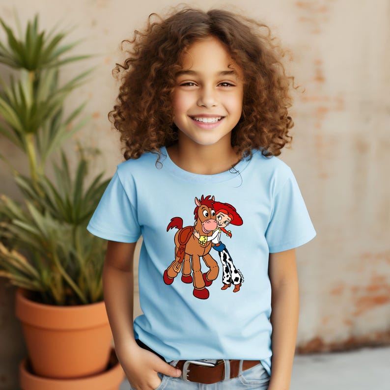 Puede incluir: Una camiseta azul claro con un dibujo animado de un caballo y una vaquera. El caballo es marr&oacute;n con una melena roja y un collar amarillo. La vaquera lleva un sombrero rojo, una camisa blanca y pantalones azules con un estampado en blanco y negro.