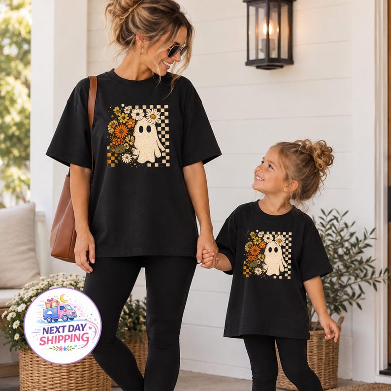 May include: Two black t-shirts featuring a graphic print of a white ghost surrounded by orange, yellow, and white flowers against a checkered background. The design includes a cluster of daisies and autumn-toned blossoms arranged beside the ghost.