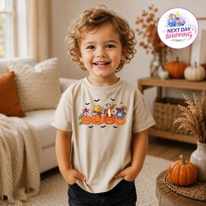 May include: A beige short-sleeve t-shirt featuring a graphic of four cartoon characters sitting inside orange jack-o'-lantern pumpkins. The design includes Piglet, Winnie the Pooh, Tigger, and Eeyore surrounded by small black bats and spiderwebs.